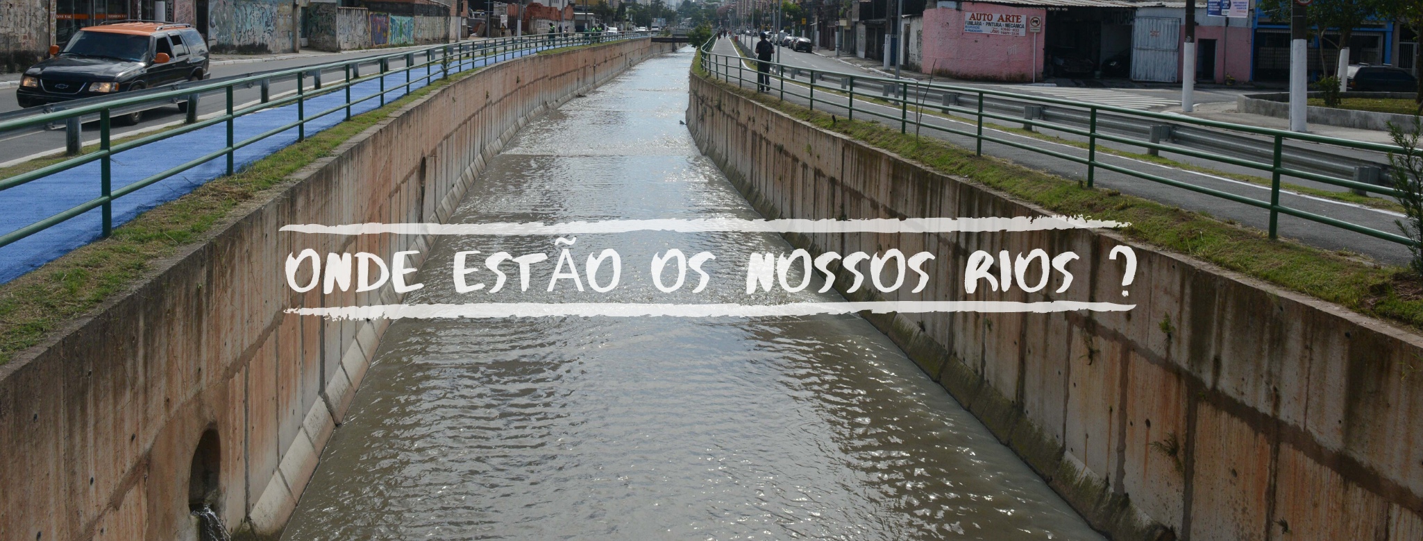 Uma foto de um rio canalizado em uma área urbana, com muros de concreto e água barrenta fluindo no centro. Há uma rua com tráfego e calçadas com grades de proteção nas laterais. No centro da imagem, sobre a água, lê-se "ONDE ESTÃO OS NOSSOS RIOS ?" em letras brancas