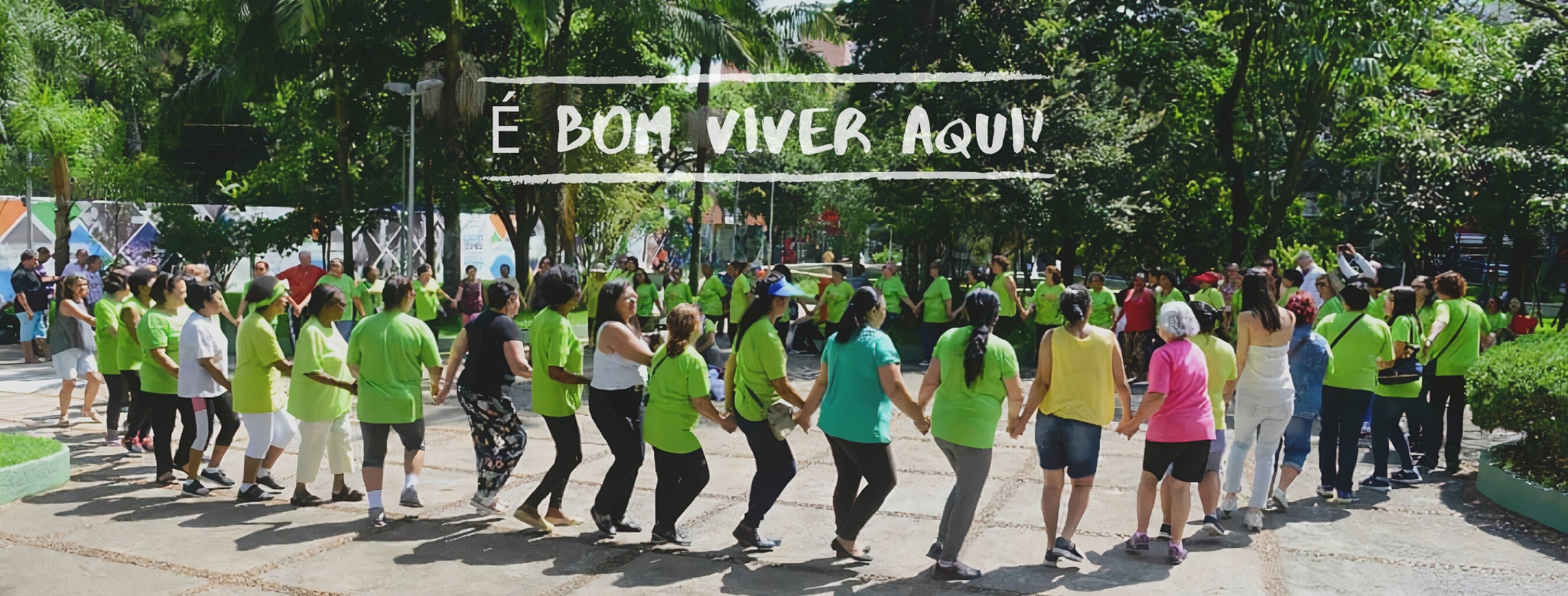 Uma foto mostra um grupo de pessoas, a maioria idosa, formando uma grande roda de mãos dadas em um parque arborizado. Ao centro, o texto "É BOM VIVER AQUI".